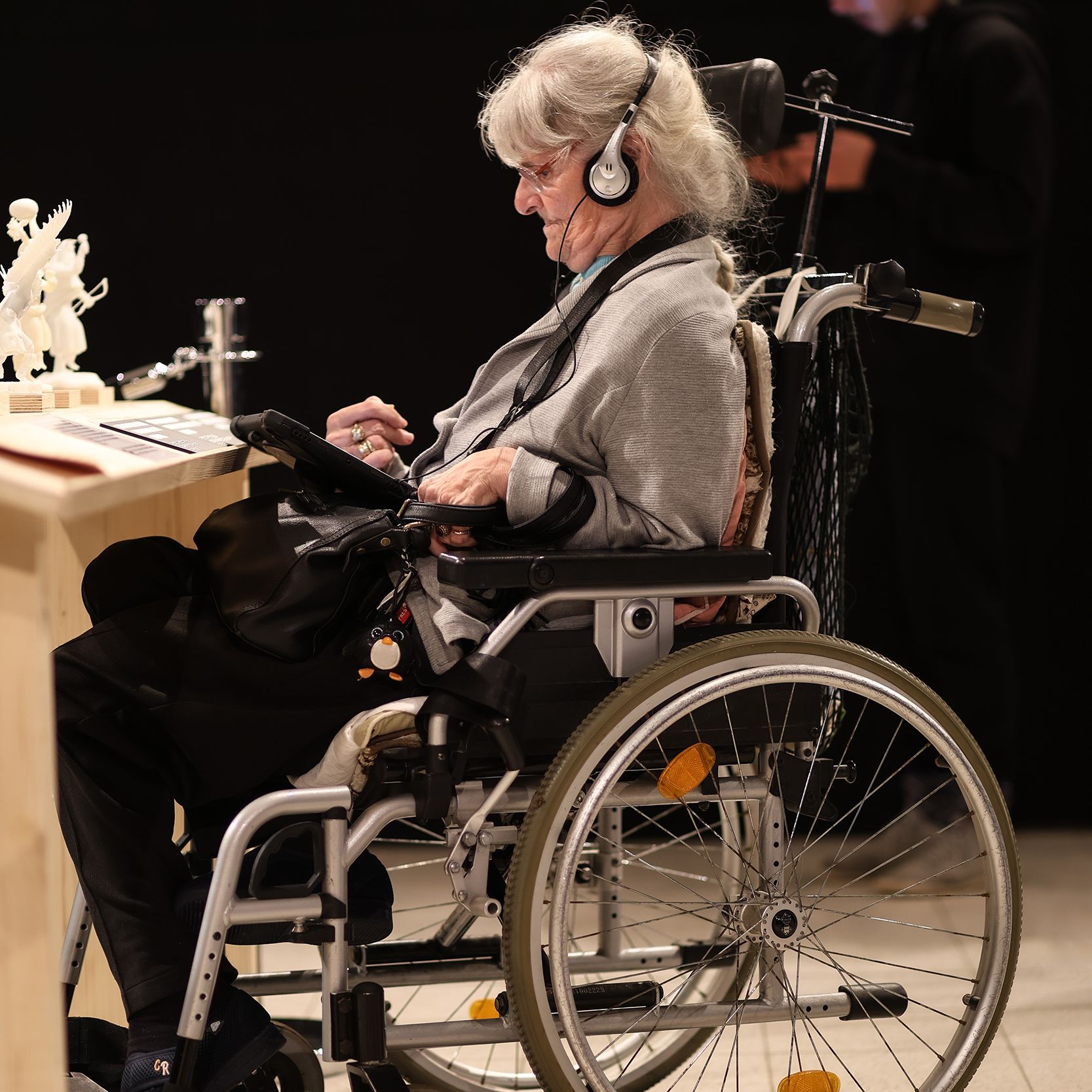 ältere Frau im Rollstuhl vor Tisch in Ausstellung. Sie hat Kopfhörer auf und schaut auf ein Tablet auf ihrem Schoß.