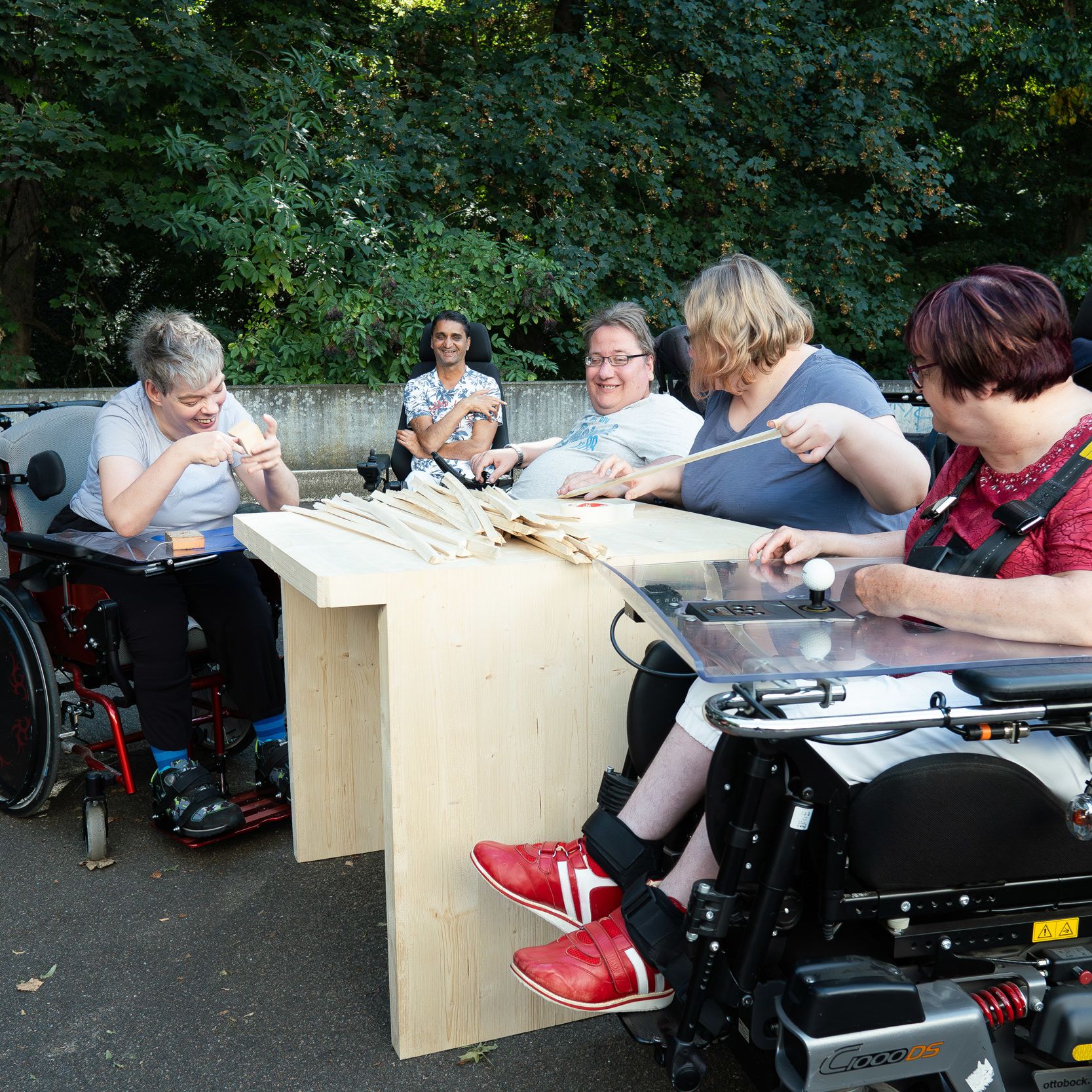 fünf Personen im Rollstuhl draußen beim Schleifen von Holzteilen, alle lachen